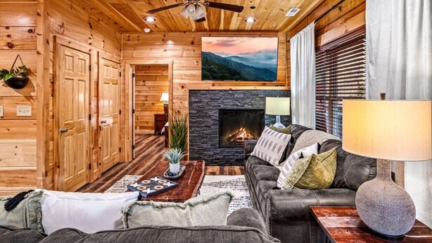 A log cabin interior with a fireplace, wooden walls, a ceiling fan, and a couch. - 5BD: Prime Pigeon Forge with 2 Hot Tubs, Pool Table, Theater, Arcade and 3 Decks (Pigeon Forge)