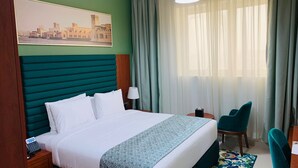 Habitación de hotel con una cama grande, un escritorio, una silla, una lámpara y vistas de la ciudad a través de cortinas translúcidas.