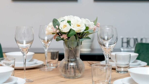 A dining table set with white plates, clear glasses, and a vase with white flowers.