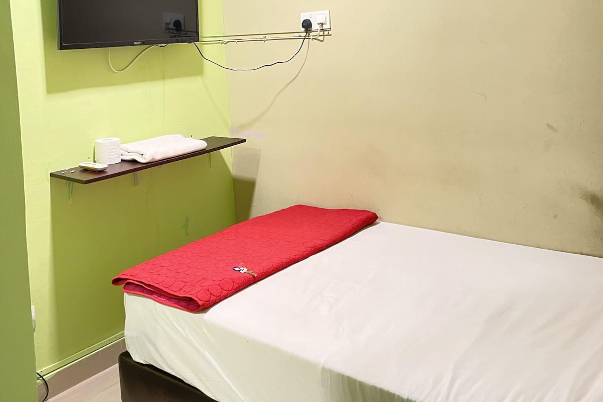 A small, single-bed room with a red blanket, a television mounted on the wall, and a shelf with toiletries.