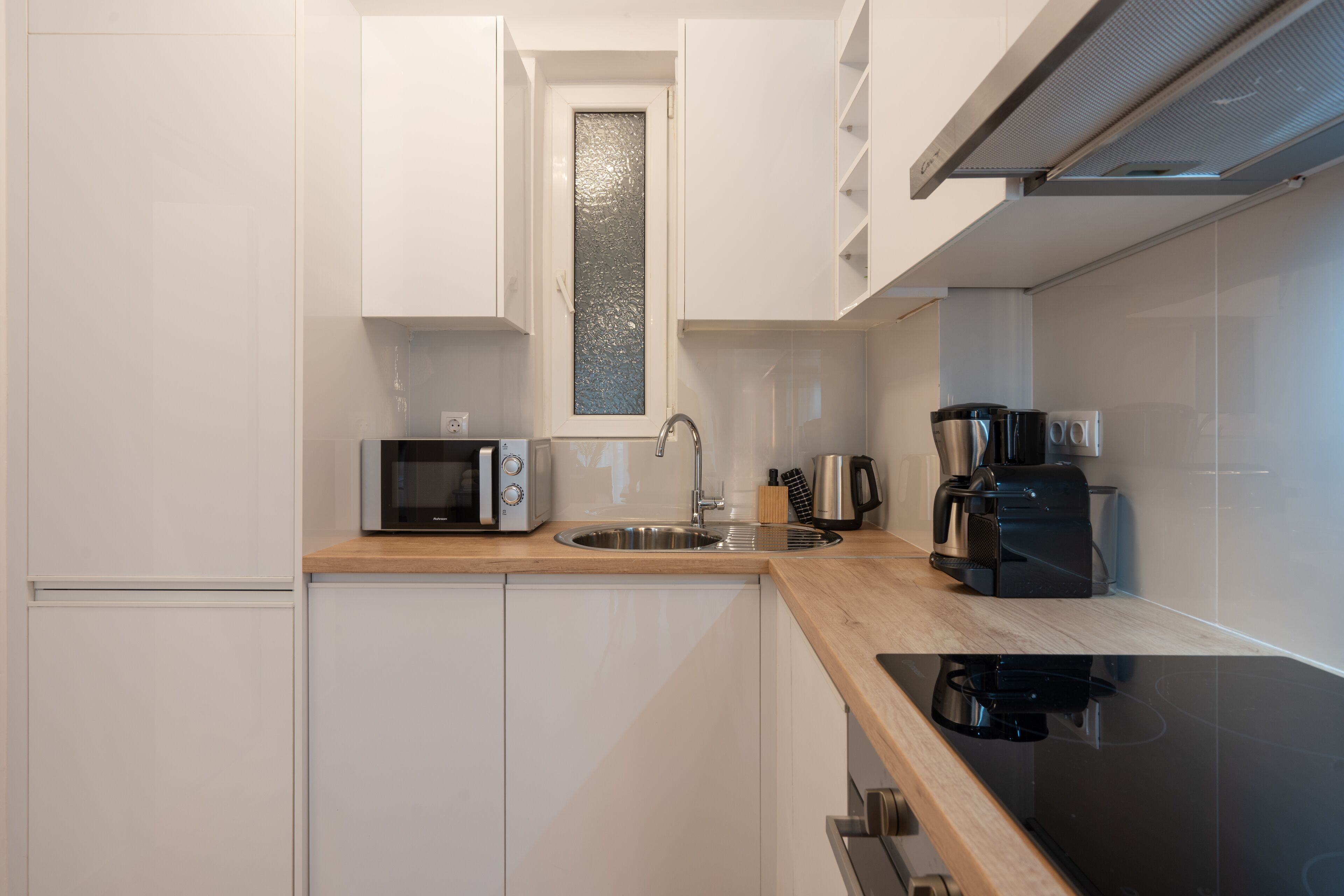 Una cocina compacta con armarios blancos, un lavamanos de acero inoxidable, un microondas, una cafetera y un tostador.