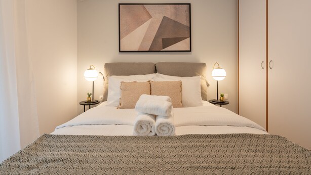 A neatly made bed with a patterned bedspread, two bedside lamps, and a geometric artwork above the bed. - Chic and Lavish Apartment in Athens (Kallithea)
