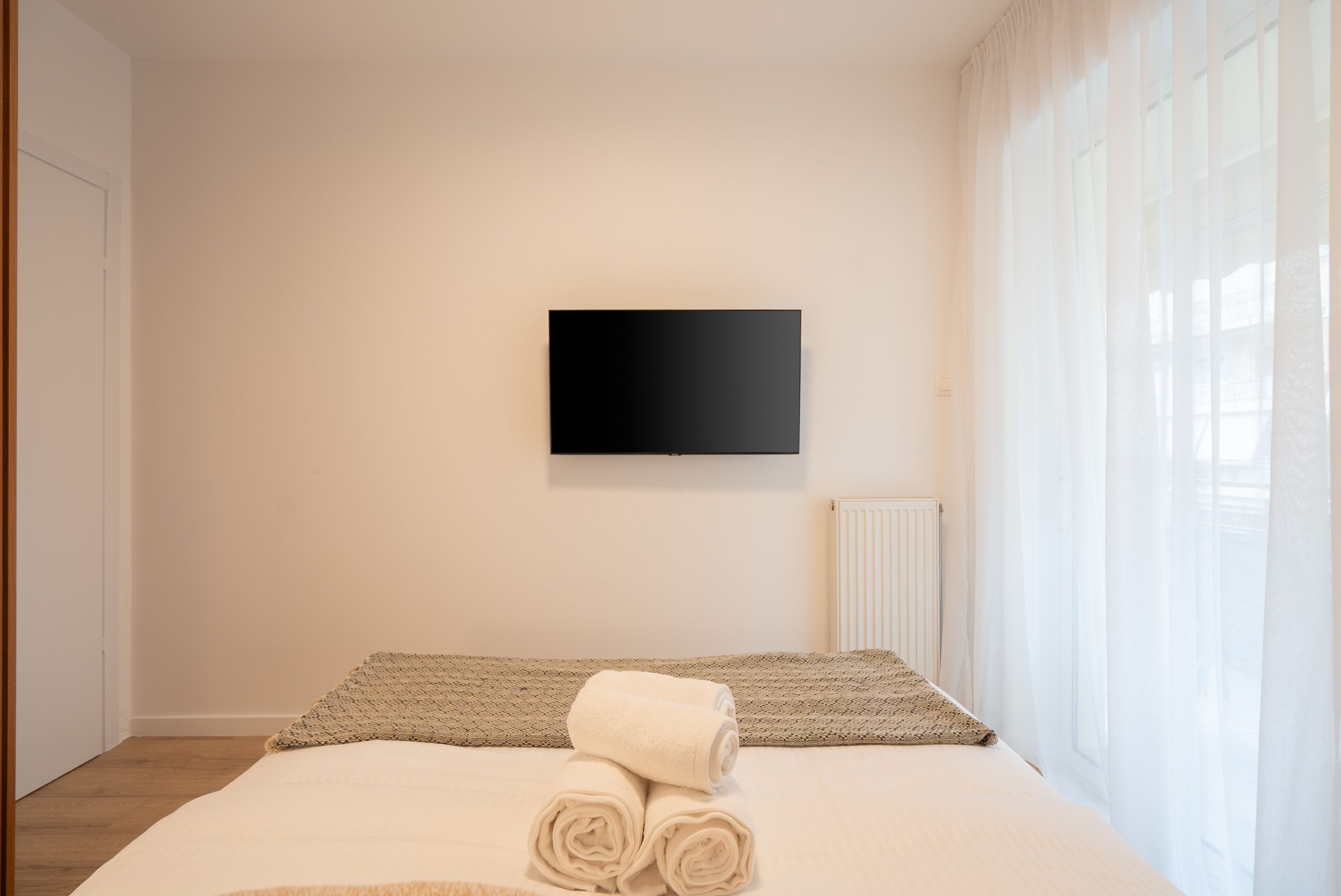 Quarto de hotel com cama, televisão de tela plana na parede e janela com cortinas leves.