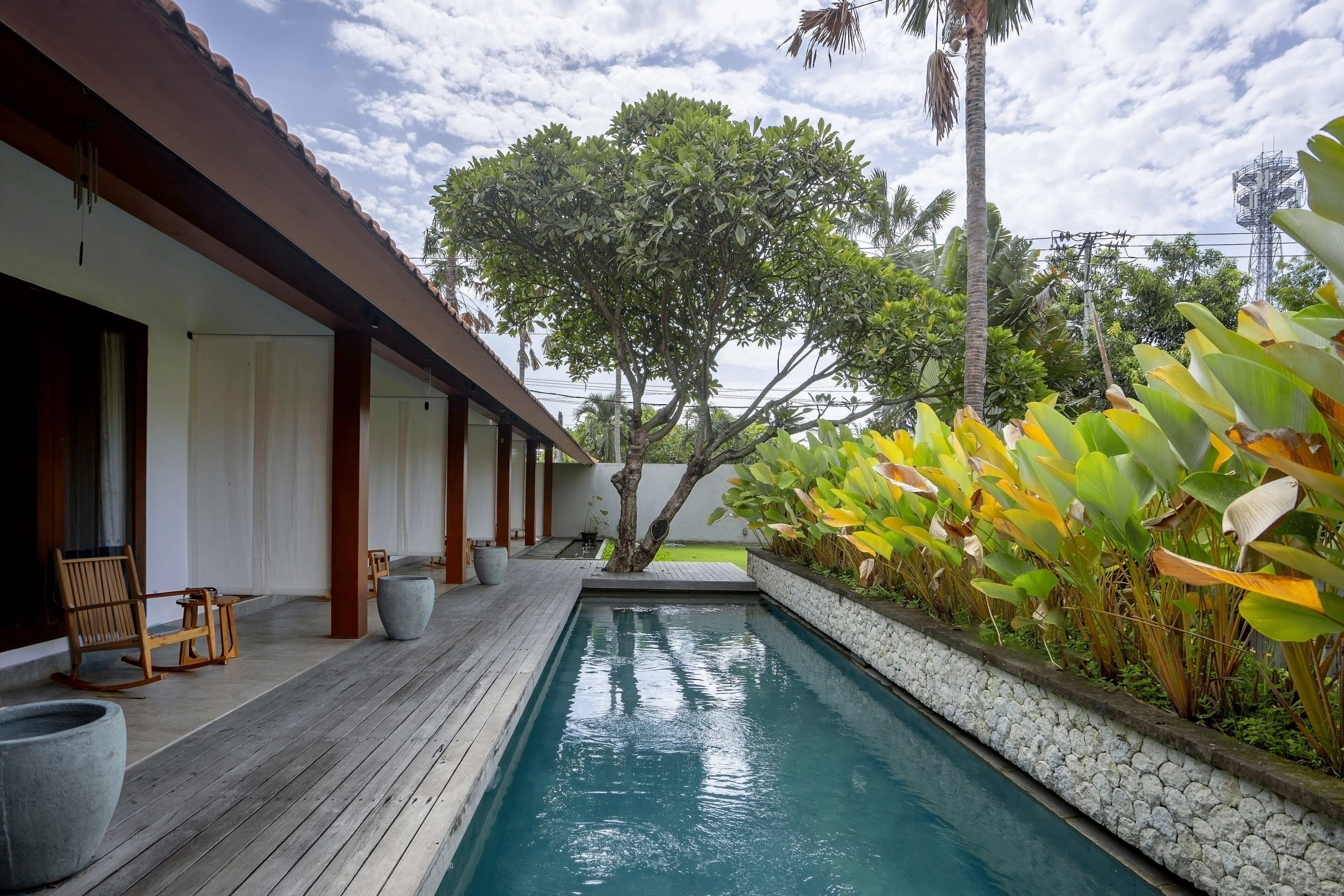 A pool area with a wooden deck, seating, and tropical plants.
