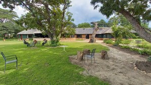 A courtyard with a thatched-roof building, a grassy lawn, and a few chairs. - ZIMPARKS BOOKINGS (Hammanskraal)