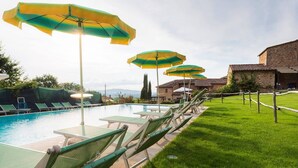 Uma área de piscina com várias espreguiçadeiras e guarda-sóis, um gramado e um edifício de pedra ao fundo.