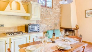 Cozinha com parede de pedra, fogão a gás, rádio antigo e mesa de jantar posta para dois, com taças de vinho e pratos.