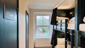 Una habitación con una cama litera, una ventana con vistas a la vegetación, un radiador y una puerta que indica "ducha".