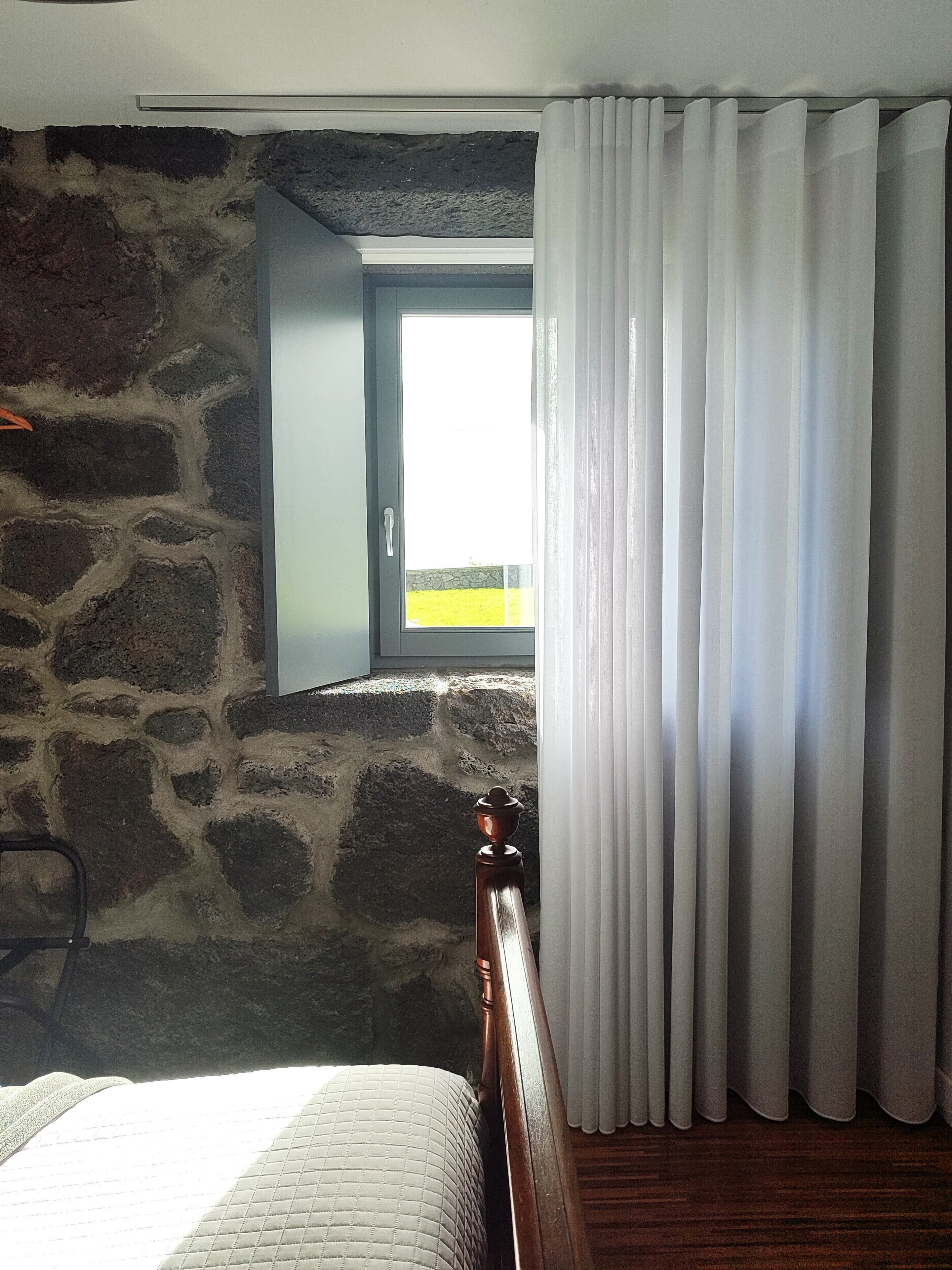 Une chambre avec un mur en pierre, un lit en bois, une fenêtre avec des rideaux et une vue sur un champ jaune.