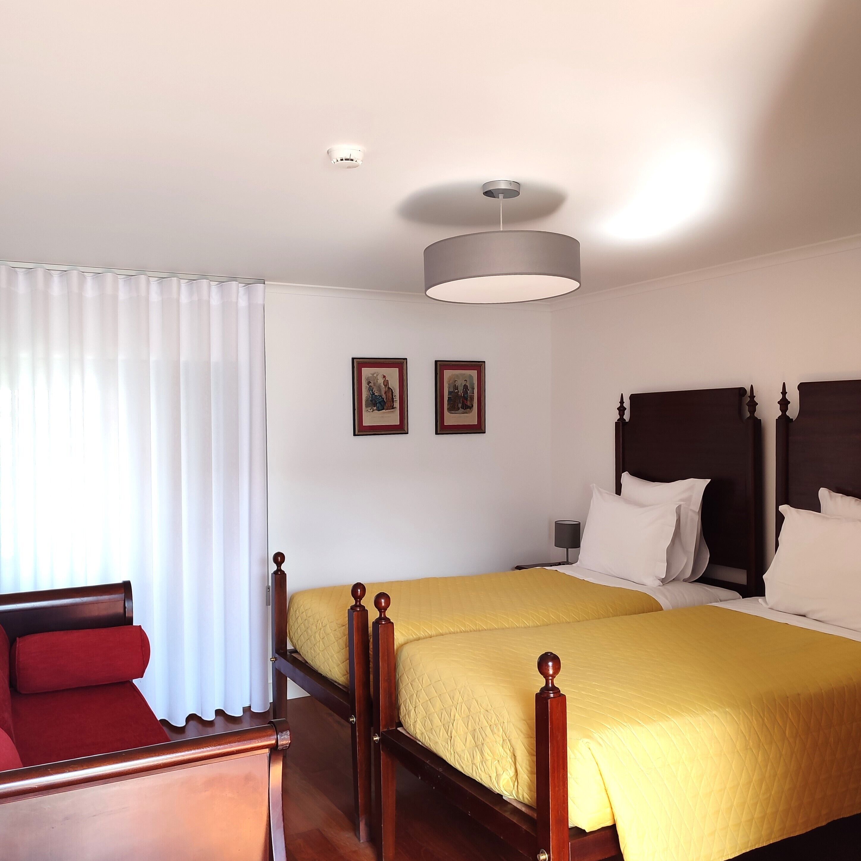 A hotel room with a double bed, a bedside table with a lamp, a window with curtains, and two framed pictures on the wall.