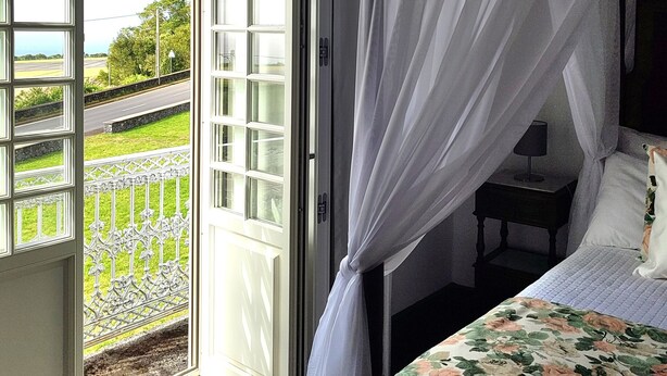 A bedroom with a bed, a nightstand, and a window with a view of a green landscape. - Quinta dos Mistérios  (Velas)