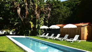 A rectangular swimming pool with white lounge chairs and umbrellas, surrounded by lush greenery and a wooden structure. - Quinta dos Mistérios  (Velas)