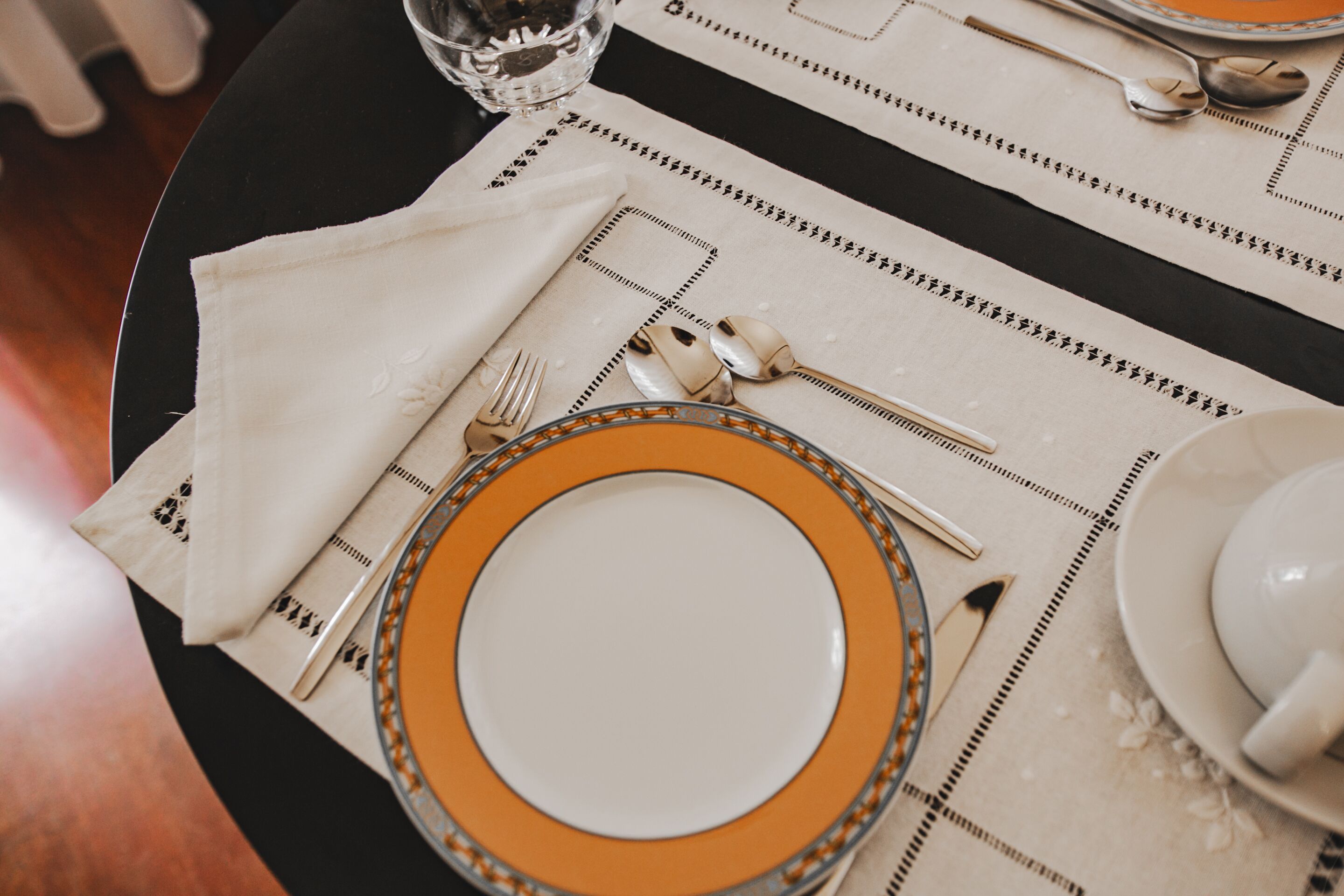 A table set with a white napkin, a plate with an orange rim, a spoon, a fork, and a glass of water.