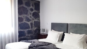 A modern bedroom with a large bed, a stone accent wall, a pendant light, and a nightstand with a lamp. - Quinta dos Mistérios  (Velas)