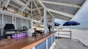 Un bar situé en bord de mer, avec un comptoir en bois, des étagères remplies de bouteilles et des parasols.