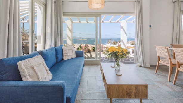 Uma sala de estar moderna com um sofá azul, uma mesa de centro de madeira e vista para o mar através de grandes janelas.