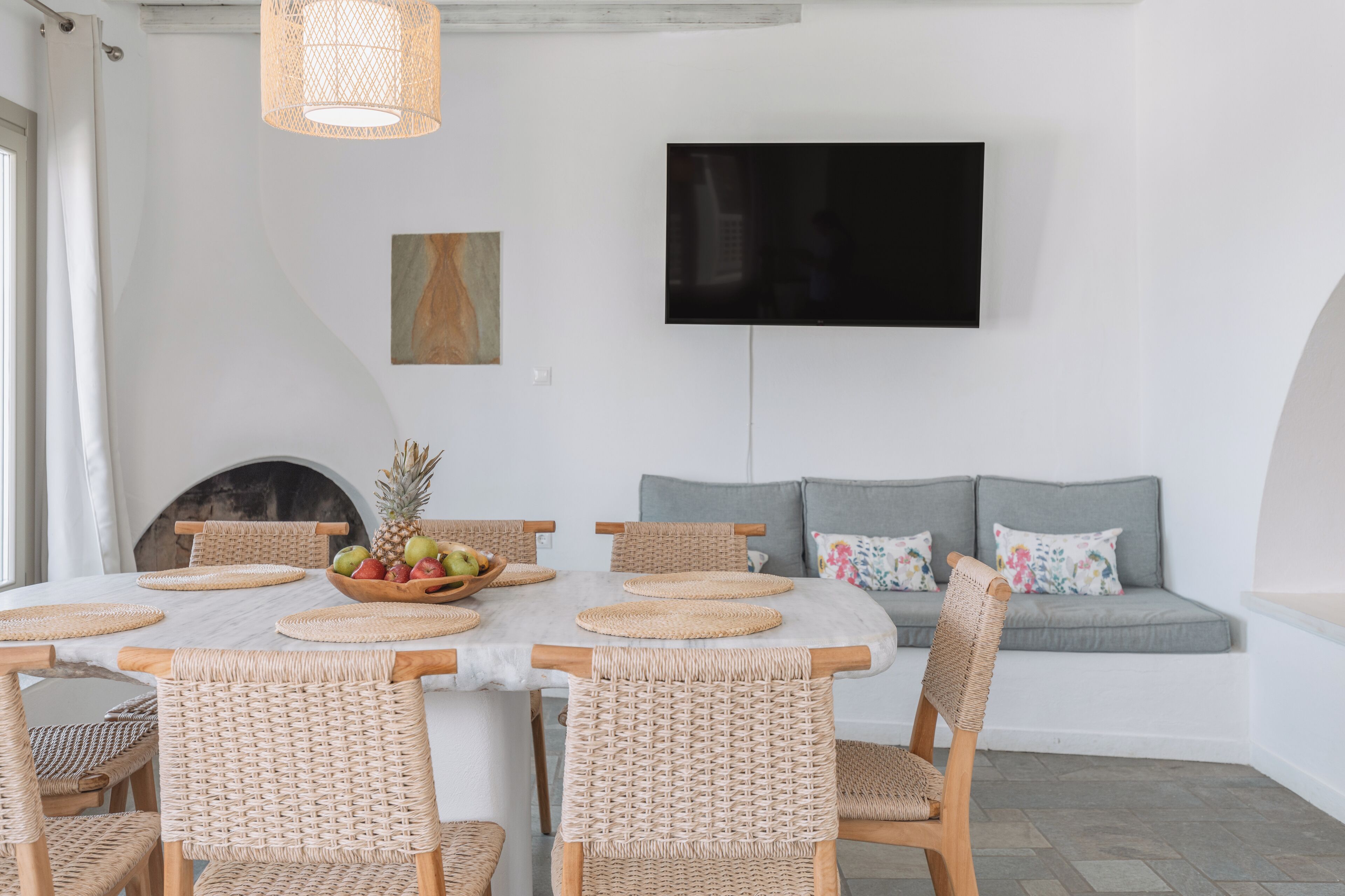 A dining area with a table set for six, surrounded by wicker chairs, a woven placemat, and a fruit bowl. A flat-screen TV is mounted on the wall above a gray sofa with floral cushions.