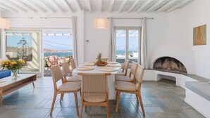 Uma sala de jantar com uma mesa redonda, cadeiras de vime, lareira e vista para o mar.