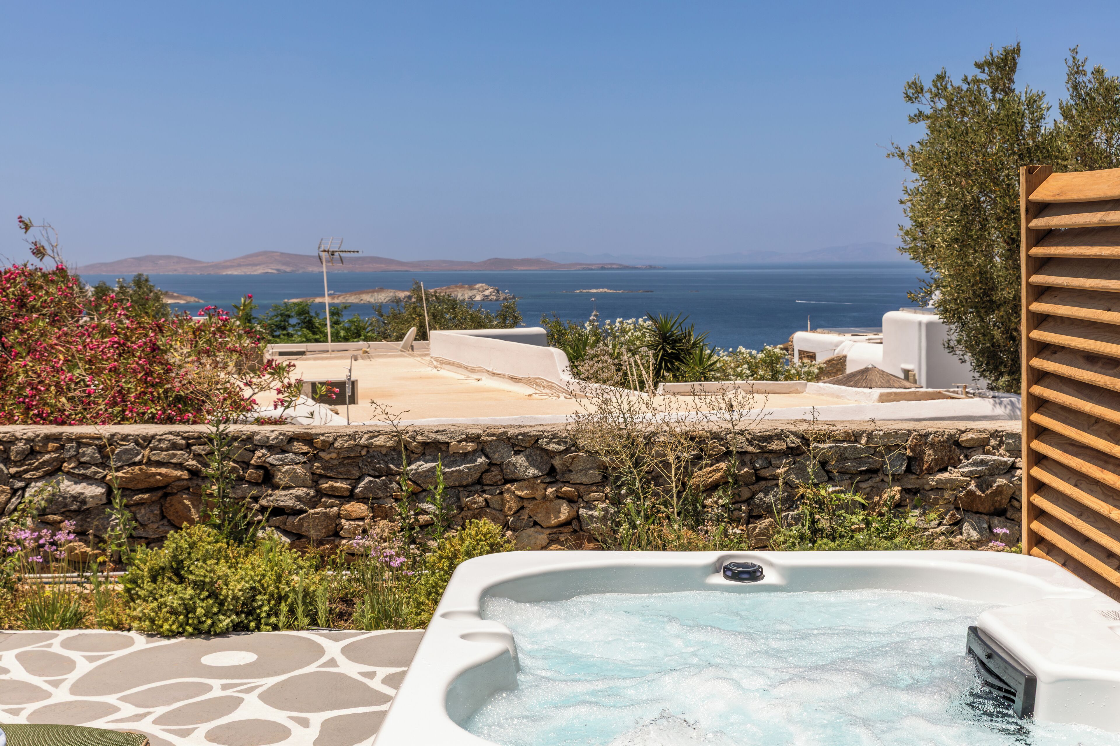 A hot tub with a stone wall and plants in the foreground, a stone pathway, and a view of the sea and mountains in the background.