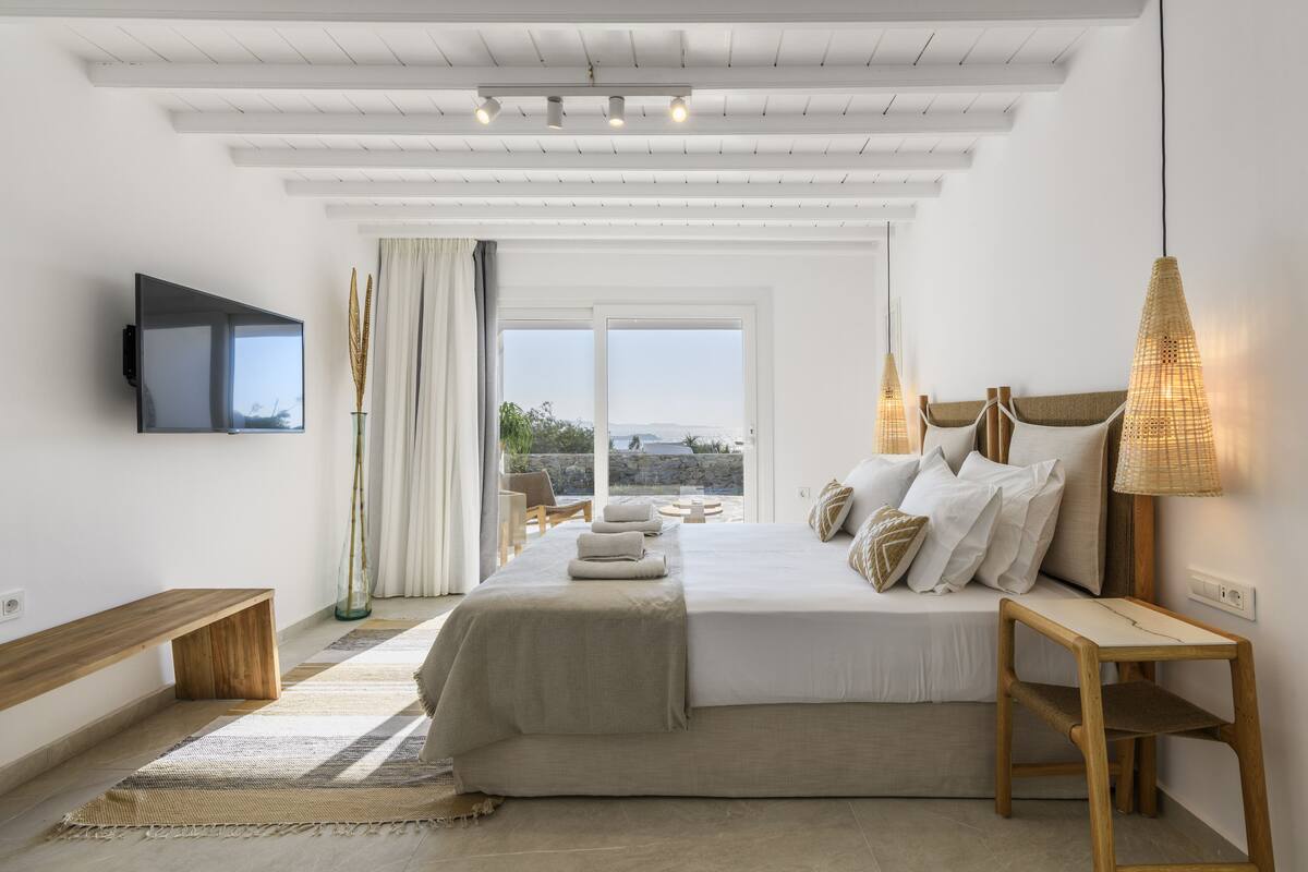 Un dormitorio con una cama grande, un televisor en la pared, un banco y vistas al exterior.