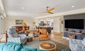 A modern living room with a sofa, armchair, and coffee table. There is a dining area with a table and chairs, and a kitchen in the background.