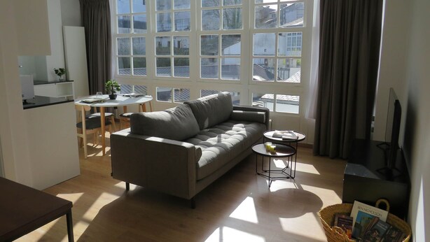 A modern living room with a grey sofa, a small round table, a dining area with a white table and chairs, and large windows with curtains.