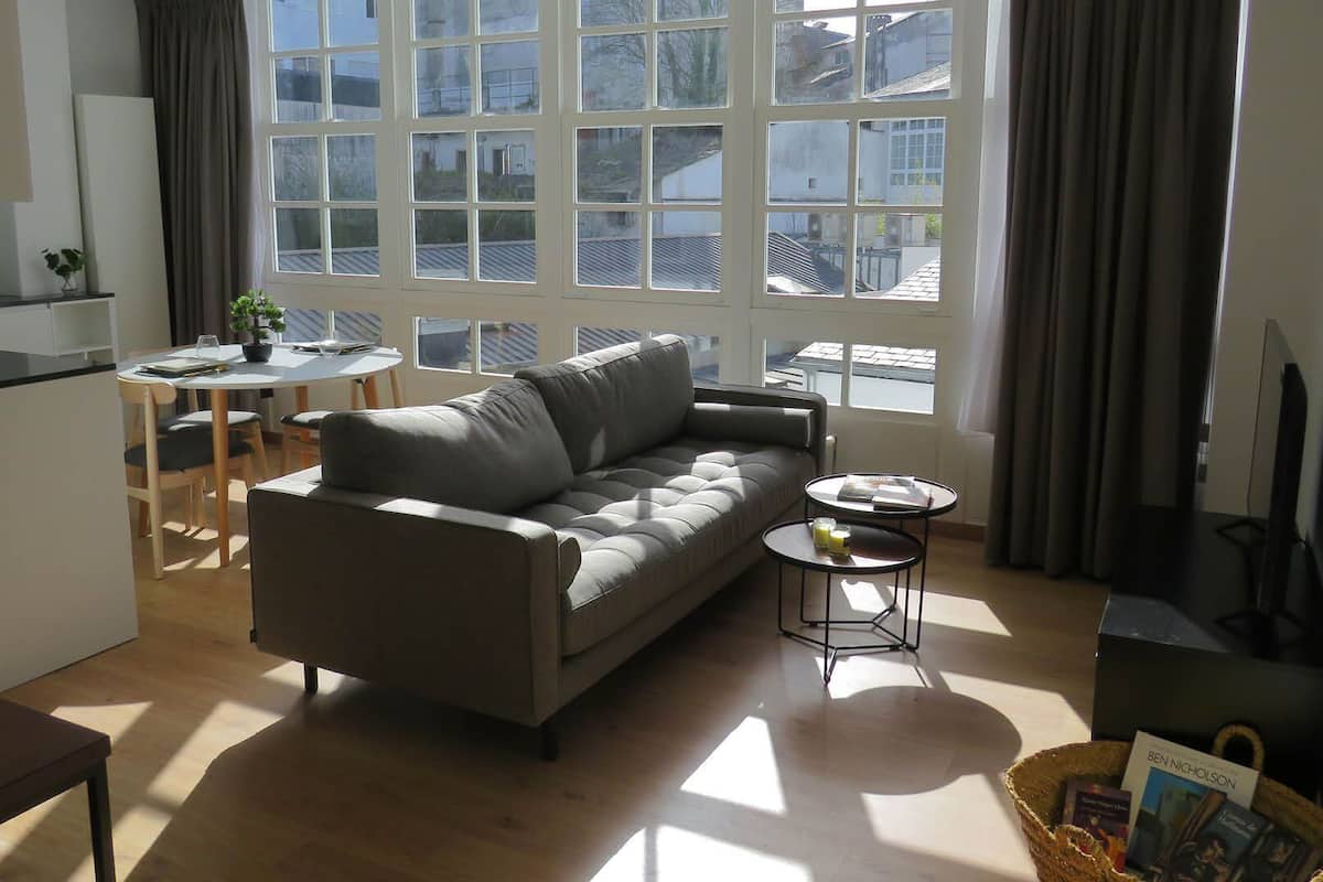 A modern living room with a grey sofa, a small round table, a dining area with a white table and chairs, and large windows with curtains.