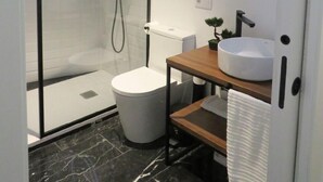 A modern bathroom with a glass-enclosed shower, a white toilet, a wooden vanity with a round mirror, and a marble floor.