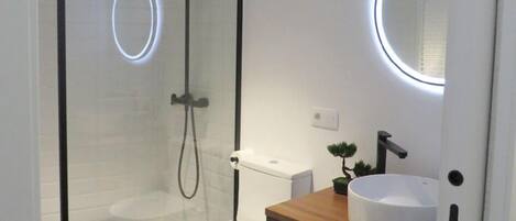 Un baño moderno con ducha envuelta en vidrio, inodoro blanco, tocador de madera con un espejo redondo y suelo de mármol.