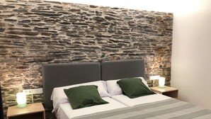 Una cama con pared de piedra de fondo, dos mesitas de noche y dos almohadas verdes.