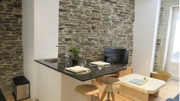 Una cocina moderna con pared de piedra, encimera negra, un lavamanos, un televisor y taburetes de madera.
