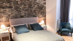 A bed with a stone wall backdrop, a nightstand with a lamp, and a chair.