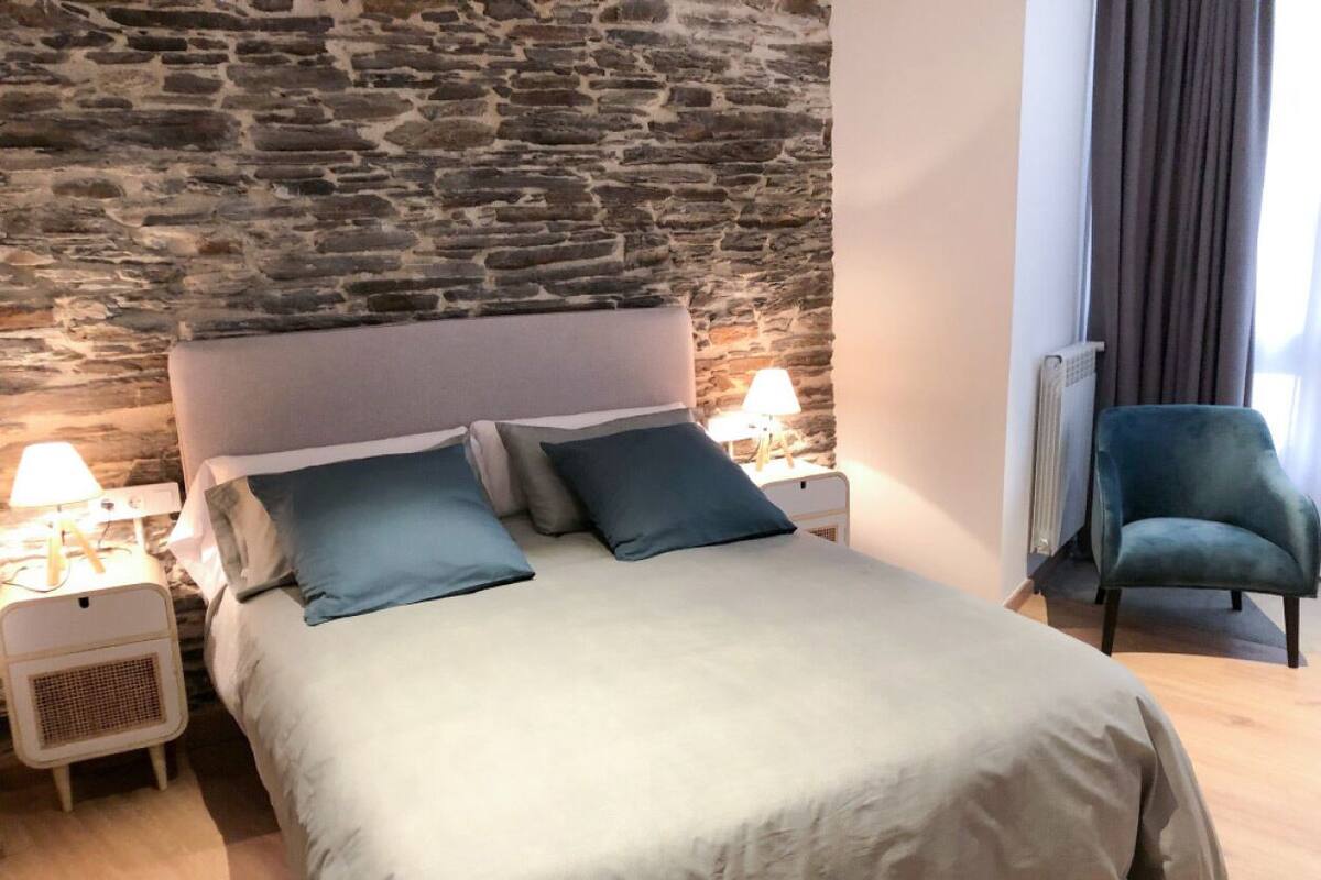 A bed with a stone wall backdrop, a nightstand with a lamp, and a chair.