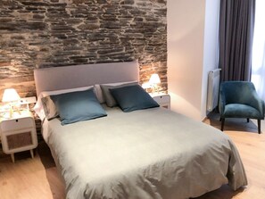 A bed with a stone wall backdrop, a nightstand with a lamp, and a chair.