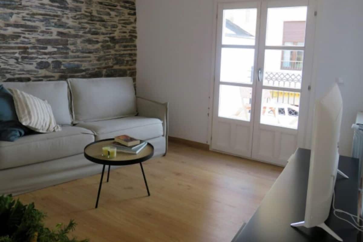A living room with a stone wall, a white sofa, a round coffee table with books, and a sliding glass door leading to an outdoor balcony.