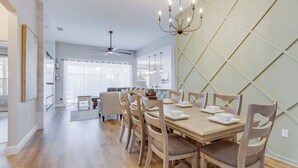 Un comedor con una mesa de madera dispuesta para cuatro personas, rodeada de sillas a juego. Se ve un ventilador de techo y una lámpara de araña encima de la mesa.