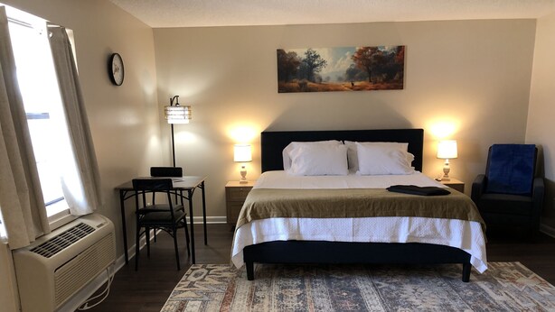 A hotel room with a bed, two bedside lamps, a desk with a chair, a rug, a painting on the wall, and a window with curtains.