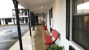 A covered walkway with benches and potted plants, leading to a building with large windows and a brick wall.