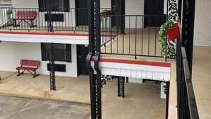 A covered outdoor area with a red bench, a black metal railing, and a white building with black shutters.