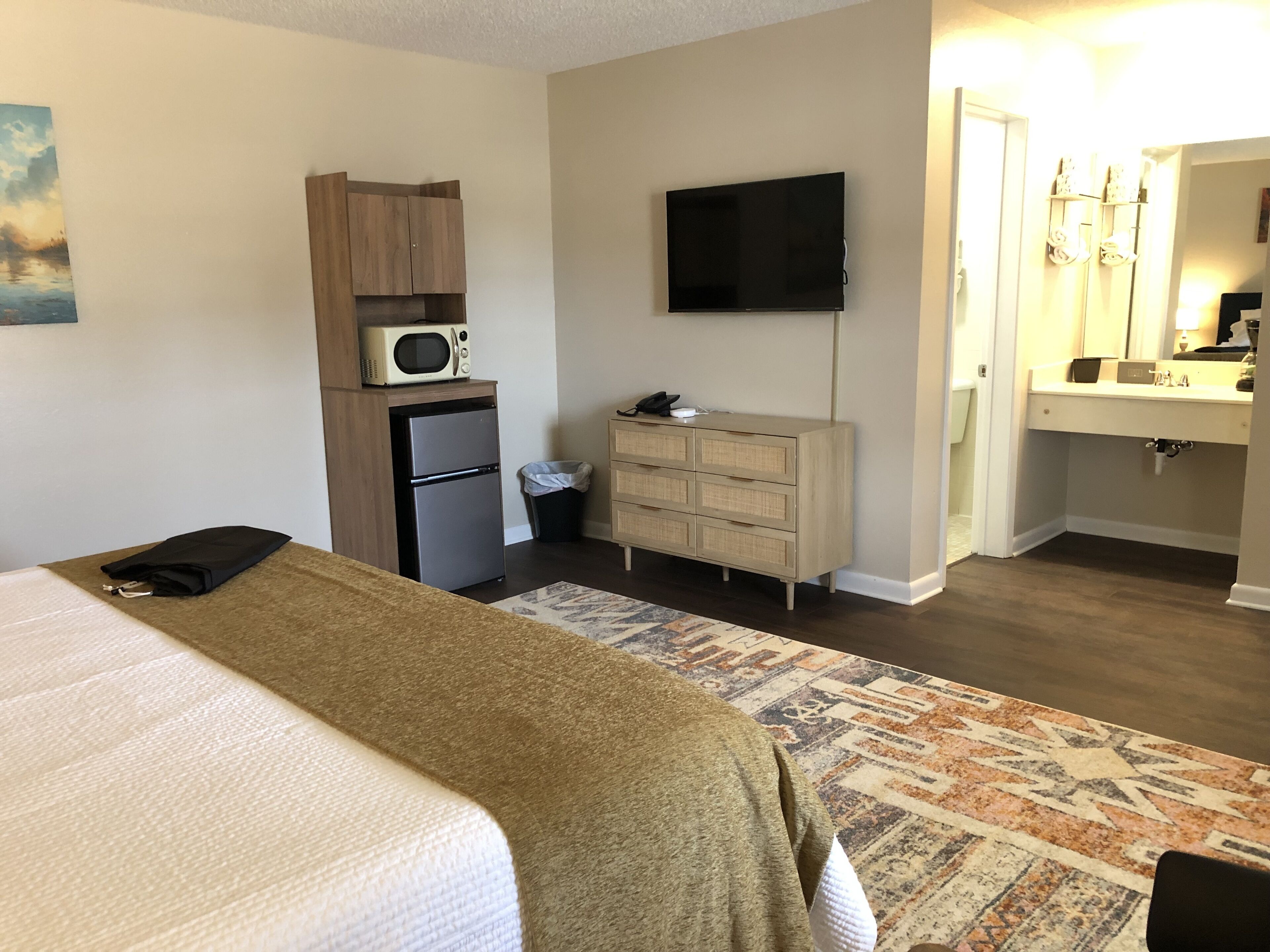 Une chambre d’hôtel comprenant un lit, une kitchenette équipée d’un four à micro-ondes et d’un réfrigérateur, une télévision, une commode et une salle de bain avec un lavabo et un miroir.