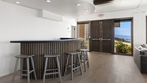 Uma área de bar moderna com balcão de madeira e quatro banquetas, vista para o mar através de grandes portas de vidro, e uma placa de saída acima das portas.