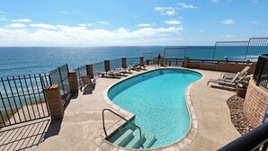 Piscina com design curvo, rodeada por espreguiçadeiras e uma cerca, com vista para o oceano.