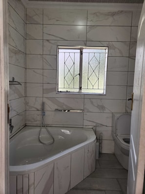 A bathroom with a marble tiled wall, a freestanding bathtub, a toilet, and a window with a diamond pattern. - Immaculate 3-bed House in Alberton Johannesburg (Alberton)