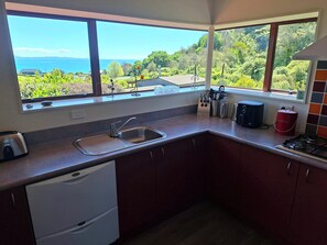 A kitchen with a view of the sea and greenery, a sink, a stove, and a dish rack. - Pohara's Kauri Lodge - Coastal Views in Golden Bay (Pōhara)