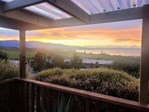 A view from a covered balcony overlooking a landscape with fields, trees, and a body of water during sunset.