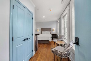 Une chambre d’hôtel avec une grande porte bleue, un lit avec une literie blanche, deux tables de chevet avec des lampes, un fauteuil en osier et une petite table avec une chaise.