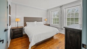 Een slaapkamer met een houten vloer, een bed met witte lakens, twee nachtkastjes met lampen en een raam met zonwering.