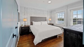 Un dormitorio con pared de ladrillo blanco, suelo de madera, cama con ropa de cama blanca, dos mesitas de noche con lámparas y una ventana con persianas.