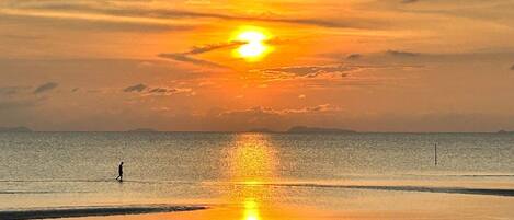 Escena de playa con un atardecer, mar tranquilo y una persona caminando por la orilla.