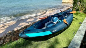 Un kayak inflable azul con una remo naranja descansando sobre una zona cubierta de hierba junto a una orilla rocosa con el océano al fondo.